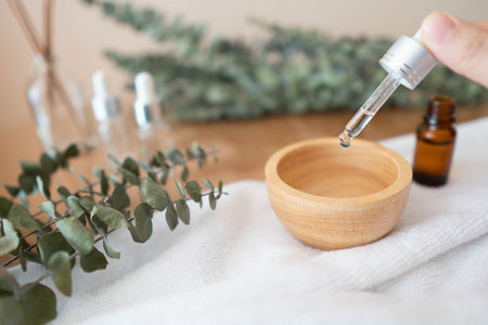 Woman's hand holding pipette and dripping eucalyptus essential oil into a bowl with natural eucalyptus leaves on wooden table. Aromatherapy, spa, skincare or herbal medicine concept.の写真素材