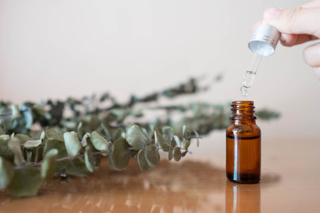 Naturalâ eucalyptusâ essentialâ oilâ forâ herbal medicine, skincare, spa or aromatherapy.â Woman's hand dripping oil into brownâ glassâ bottleâ withâ organic eucalyptusâ leaves onâ wooden table.の写真素材