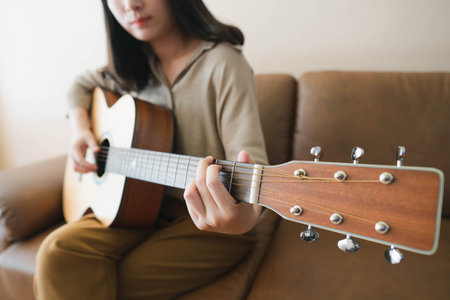 Beautiful asian woman playing acoustic guitar while sitting on a couch at home. Hobbies and leisure activities concept. Selective focus.の写真素材