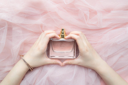 Women hands making a heart shape with perfume bottle on pink folded fabric background. Scent fragrance cosmetic beauty product.の写真素材