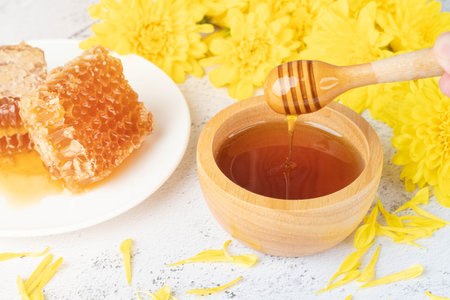 Honey dripping from honey dipper in wooden bowl with fresh honeycomb and wild flower on white table.の写真素材