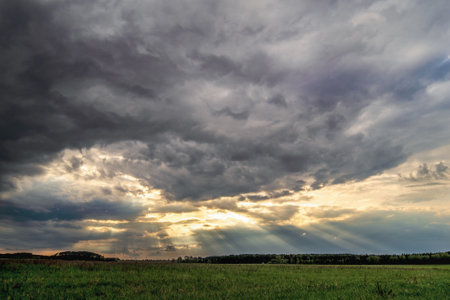 Dramatic landscape with dark storm clouds and sun rays breaking through gaps in the sky. The light illuminates the green field and horizonの写真素材