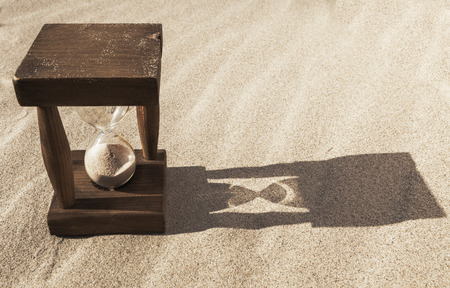 Old wooden hourglass in the sand with morning long shadow
