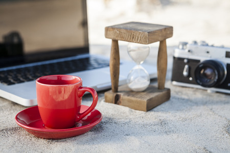 Red Coffe Mug and Vintage hourglass with silver laptop and old camera in the sand at the beachの写真素材