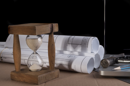 Architectural plans rolled with silver laptop hammer and a pencil and old wood hourglassの写真素材