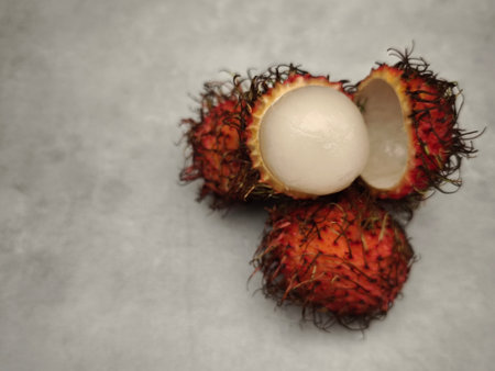 Ripe rambutan fruit open revealing its juicy white flesh, resting on a textured gray surfaceの写真素材