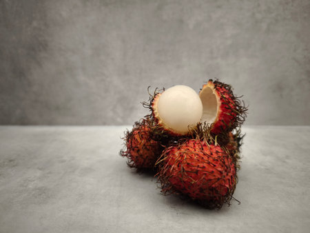 Ripe rambutan fruit, partially peeled to reveal its white flesh, sits on a gray table against a textured gray backdropの写真素材