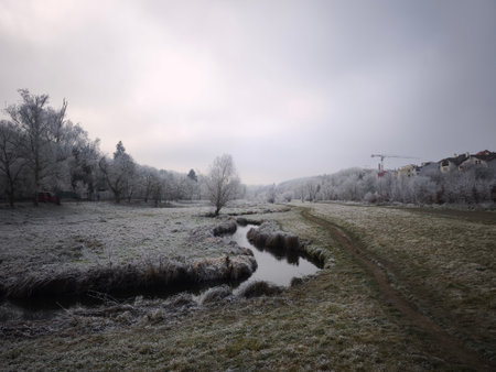 Scenic winter landscape showcasing a frozen creek winding through frosty fields in Prague 9,...