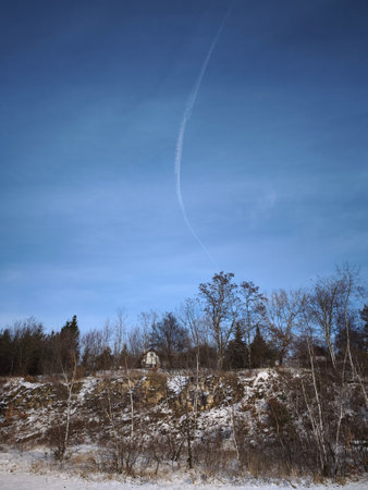 White contrail curving across blue sky over winter landscape with snowy trees and rocky cliff,...
