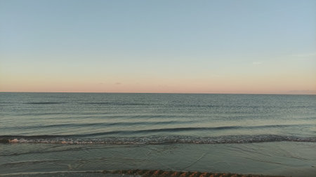 Scene of waves washing ashore as the sun sets, painting the sky with soft hues of pink and orange in...