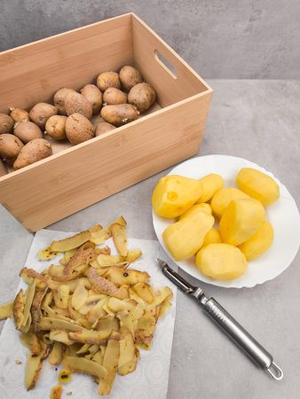 Peeled and unpeeled potatoes, along with their peels and a peeler, arranged on a kitchen counter, preparing for delicious cookingの写真素材