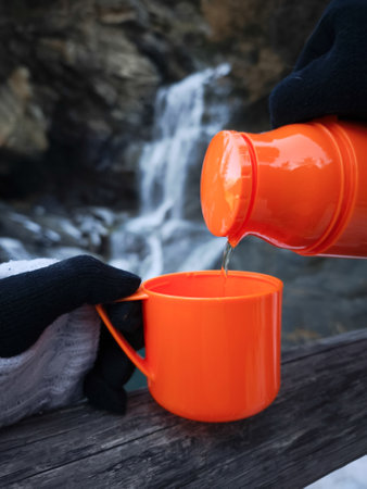 Gloved hands pouring a hot beverage from a thermos into a cup, set against the backdrop of a waterfall in aosta, italy during november Cascate di Lillazの写真素材