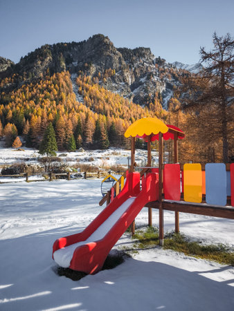 Brightly colored playground equipment stands against a backdrop of snow covered ground and autumnal trees in aosta, italy, surrounded by majestic mountains Frazionne Lillazの写真素材