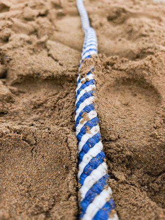 Close up of a blue and white rope partially submerged in the sand on a beach, creating a contrast of...