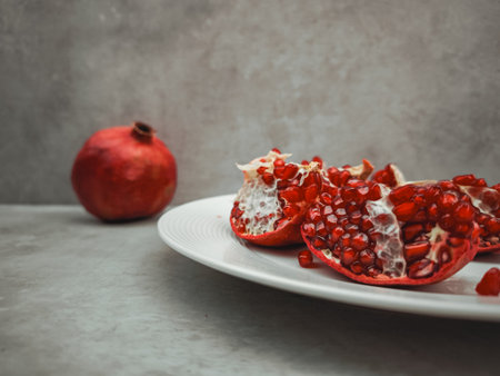 Displaying ripe pomegranate pieces on a white plate, symbolizing rosh hashanah and themes of rebirth, love, and abundance, with a whole pomegranate in the backgroundの写真素材