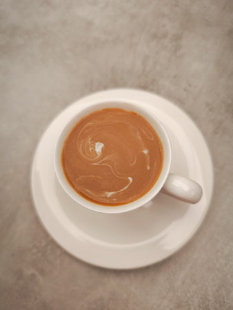 White cup filled with creamy coffee creating swirling patterns, resting on a matching saucer against a textured gray backdrop, offering a cozy and inviting atmosphereの写真素材