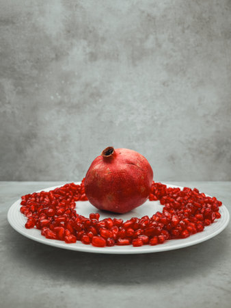 Whole pomegranate sits on a white plate surrounded by a ring of its vibrant red seeds, creating a symbolic image of love, rebirth, and traditionsの写真素材