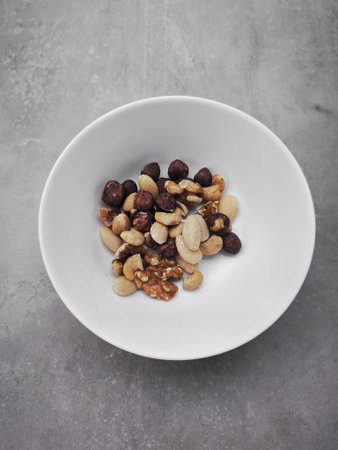 Small white bowl sits on a gray surface, containing a variety of mixed nuts, including walnuts, almonds, hazelnuts, and cashews, offering a healthy and nutritious snackの写真素材