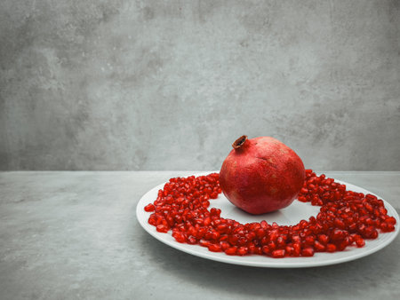 Whole pomegranate sits on a white plate surrounded by a ring of its vibrant red seeds, creating a symbolic representation of rosh hashanah against a textured gray backgroundの写真素材