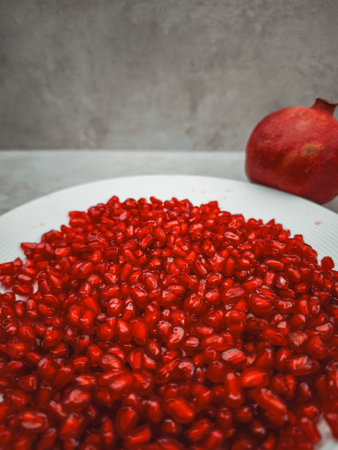 Fresh pomegranate seeds arranged on a white plate, beside a whole pomegranate, represent themes of rosh hashanah, love, rebirth, and abundance in Greek mythology and other culturesの写真素材