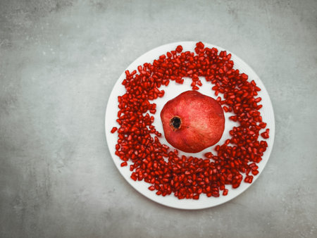Whole pomegranate resting on a white plate, surrounded by vibrant red seeds, creating a visually appealing still life symbolizing love, rebirth, and traditionの写真素材
