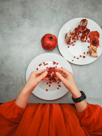 Woman wearing an orange dress. Opening a pomegranate fruit over a white plate. Symbolizing prosperity and abundance during rosh hashanah. The jewish new year. With other pomegranates on a gray tableの写真素材