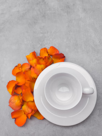 Empty white coffee cup sits on a matching saucer, surrounded by vibrant orange rose petals, creating a serene and romantic autumnal scene on a textured gray backgroundの写真素材