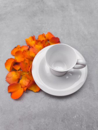 Empty white coffee cup sits on a matching saucer, accompanied by vibrant orange flower petals scattered on a gray surface, creating a serene autumnal still lifeの写真素材
