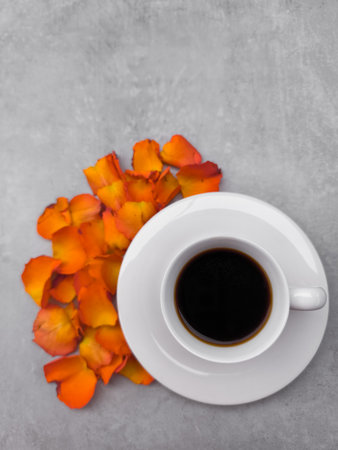 White coffee cup standing on a saucer with orange rose petals scattered around, creating a cozy and romantic autumn atmosphere on a gray background with copy spaceの写真素材