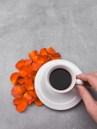 Hand holding a white coffee cup filled with rich black coffee, positioned next to a vibrant pile of orange flower petals on a soft gray background, offering ample copy spaceの写真素材