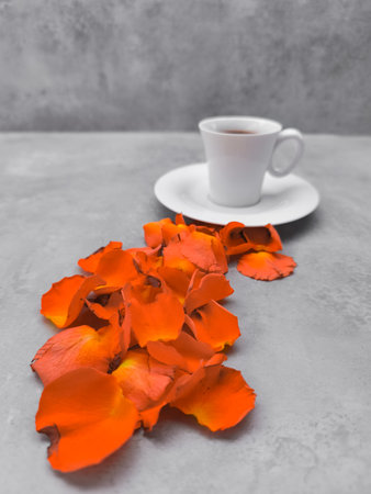 White cup filled with coffee sits on a saucer on a gray table, with a scattering of vibrant orange rose petals cascading from beneath the cup, creating a warm and inviting autumnal sceneの写真素材