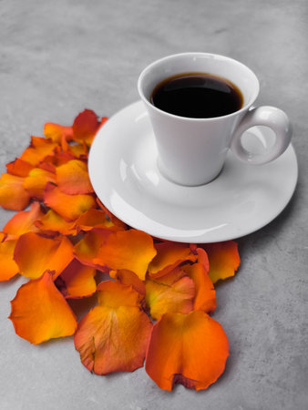 White cup of coffee sits on a white saucer on a gray table, surrounded by vibrant orange rose petals, creating a warm and inviting autumnal sceneの写真素材
