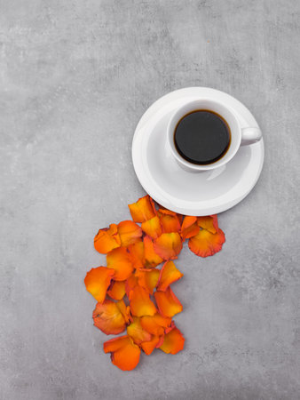 White cup of coffee sits on a white saucer on a gray background, with orange rose petals scattered around it, creating a cozy and inviting atmosphereの写真素材