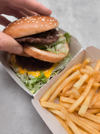 Hand holding a double cheeseburger with french fries in a cardboard container