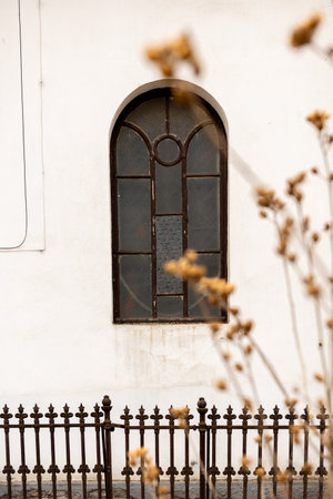 vintage brown wooden window in white wall of old church, metallic fence and gateの写真素材