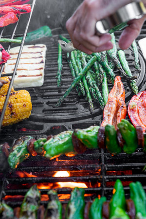Grilling corn cobs, paprika, padron, asparagus and cheese. Meat tongs in human hands.