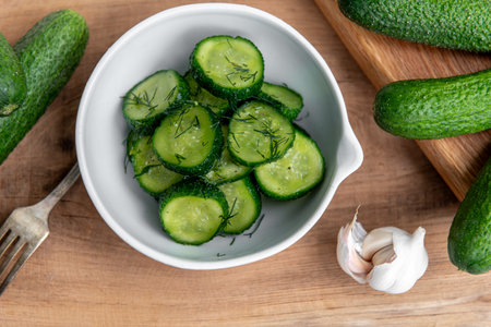 Organic cucumber slices in a ceramic white bowl on a wooden table. Fresh vegetables and dill, garlic on textured wooden table. Salad.の写真素材