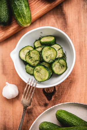 Organic cucumber slices in a ceramic white bowl on a wooden table. Fresh vegetables and dill, garlic on textured wooden table. Salad.の写真素材