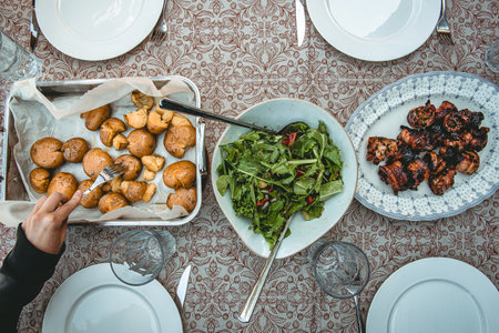 Top view of a dining table set with homemade dishes, including a bowl of fresh green salad, roasted potatoes, and grilled pork belly skewers. Rustic family-style meal served on a patterned tablecloth.の写真素材