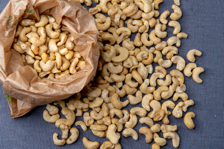 Paper bag filled with roasted cashew nuts placed on a textured gray surface, with a few nuts scattered around for a natural, appetizing look.の写真素材