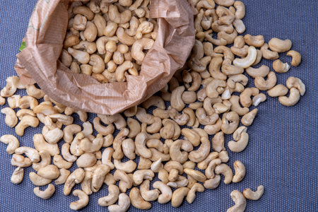 Paper bag filled with roasted cashew nuts placed on a textured gray surface, with a few nuts scattered around for a natural, appetizing look.の写真素材