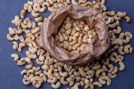 Paper bag filled with roasted cashew nuts placed on a textured gray surface, with a few nuts scattered around for a natural, appetizing look.の写真素材