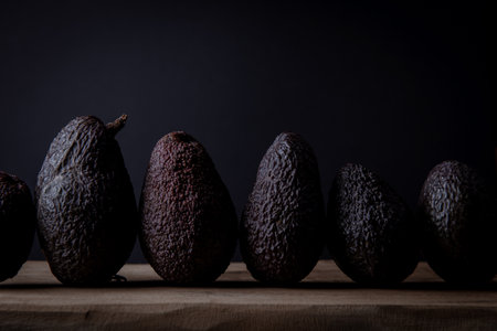Dark, ripe avocados lined up on a wooden surface with a black background, captured in soft moody...