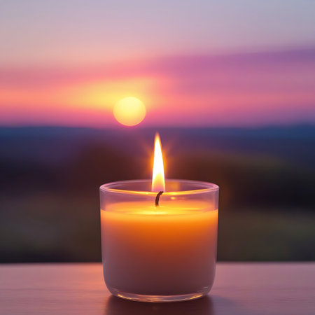 A candle with a vibrant orange and yellow flame burns in a clear glass jar. The background is a soft, out-of-focus sunset with a prominent orange sun and purple and pink sky. The image evokes a sense of calm and warmth.の素材