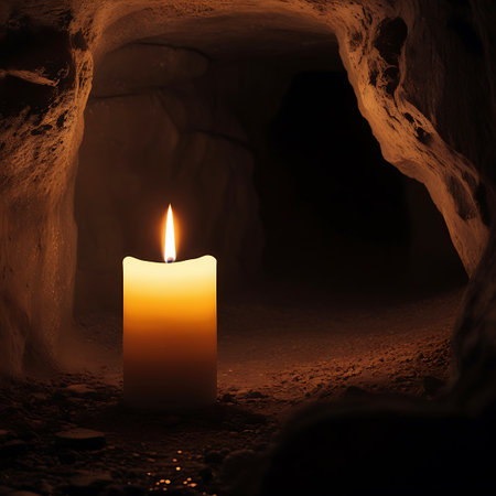 A single lit candle with a bright yellow and orange flame stands on the rocky, uneven floor of a dark cave. The rough, textured walls of the cave are illuminated by the candle's glow, creating a sense of mystery and solitude.の素材