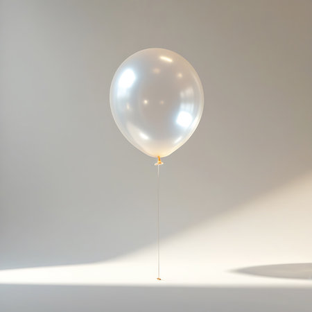 A single pearlescent white balloon floats gracefully against a softly lit white background. The balloon has a smooth, reflective surface with highlights that suggest a gentle glow. A thin golden string is attached to the bottom, trailing downwards. The lighting creates a subtle shadow on the surface below, adding depth to the minimalist composition.の素材