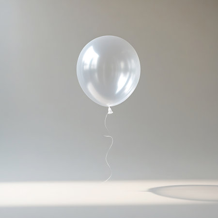 A single white balloon is presented against a light gray studio background. The balloon's surface is smooth and highly reflective, catching the light with bright highlights. A thin, delicate white string is attached to the balloon and hangs down. The lighting is soft, creating a gentle shadow on the surface beneath the balloon.の素材