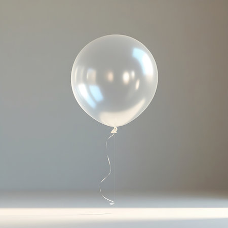 A white balloon is depicted floating in a neutral studio setting. It has a smooth, reflective surface with bright highlights. A delicate white ribbon is attached to the balloon's knot and curls gently downwards. The lighting is soft, casting a subtle shadow on the surface below, emphasizing the balloon's form.の素材