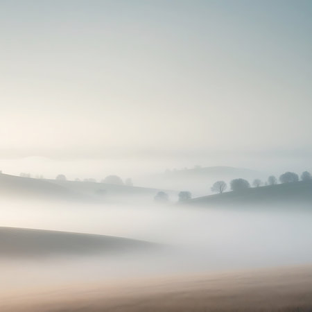 A serene landscape unfolds with rolling hills partially obscured by a thick layer of mist. Silhouetted trees dot the crests of the hills, their forms softened by the atmospheric haze. The sky above is a pale, diffused light, contributing to the overall tranquil and ethereal mood of the scene. The gentle curves of the land create a sense of depth and vastness.の素材