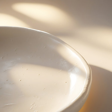 A close-up shot of the curved edge of a smooth, creamy bowl. Soft sunlight illuminates the surface, casting gentle shadows that highlight its subtle texture and form. The overall impression is minimalist and elegant.の素材